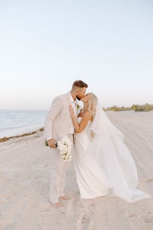 A romantic beach wedding photoshoot with a couple in white attire posing on sandy shores at sunset.
