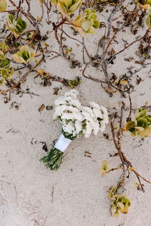 A white floral bridal bouquet lies on a textured beige surface with yellow blooms and twisting vines in the background.
