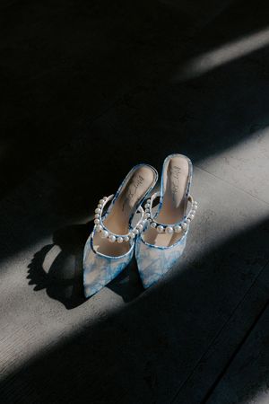 Elegant light blue embellished bridal shoes cast dramatic shadows on dark wooden floor in artistic lighting.