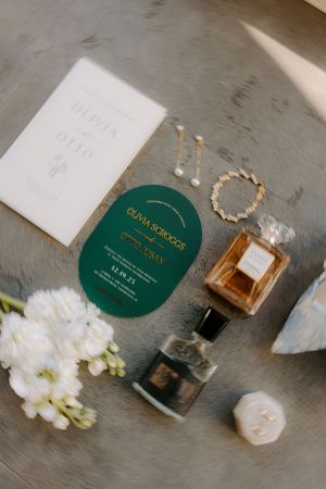 Elegant wedding details featuring perfume bottles, white flowers, and green invitation cards arranged on a gray surface.