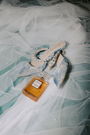 A perfume bottle and elegant blue heels arranged on delicate white tulle fabric create a romantic bridal composition.