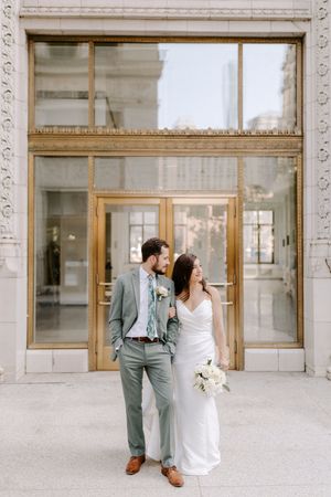 A couple stands together outside an elegant building entrance with ornate architectural details and brass doors.