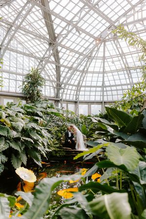 Bride and groom share a romantic moment in a lush botanical greenhouse with a stunning glass ceiling.