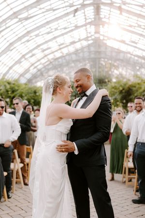 A bride and groom share a romantic first dance in a sunlit greenhouse wedding venue surrounded by guests.