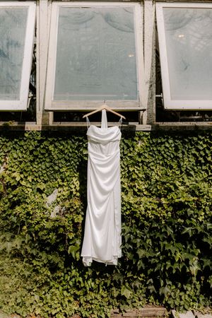 White satin wedding dress hanging on wooden hanger against ivy-covered wall with vintage windows.