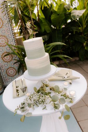 Elegant three-tier sage green wedding cake on white pedestal table with floral decor and candles outdoors.