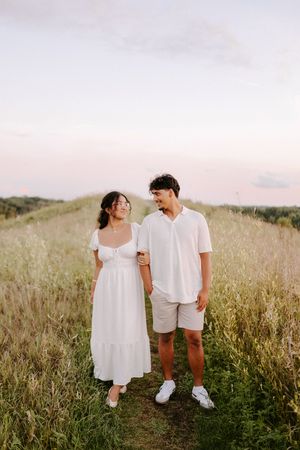 A couple in white outfits shares a tender moment while walking through a sun-lit meadow of tall grass.