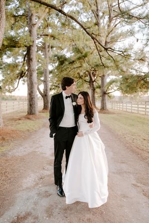 A couple in wedding attire stands together on a tree-lined dirt path with white fencing in the background.