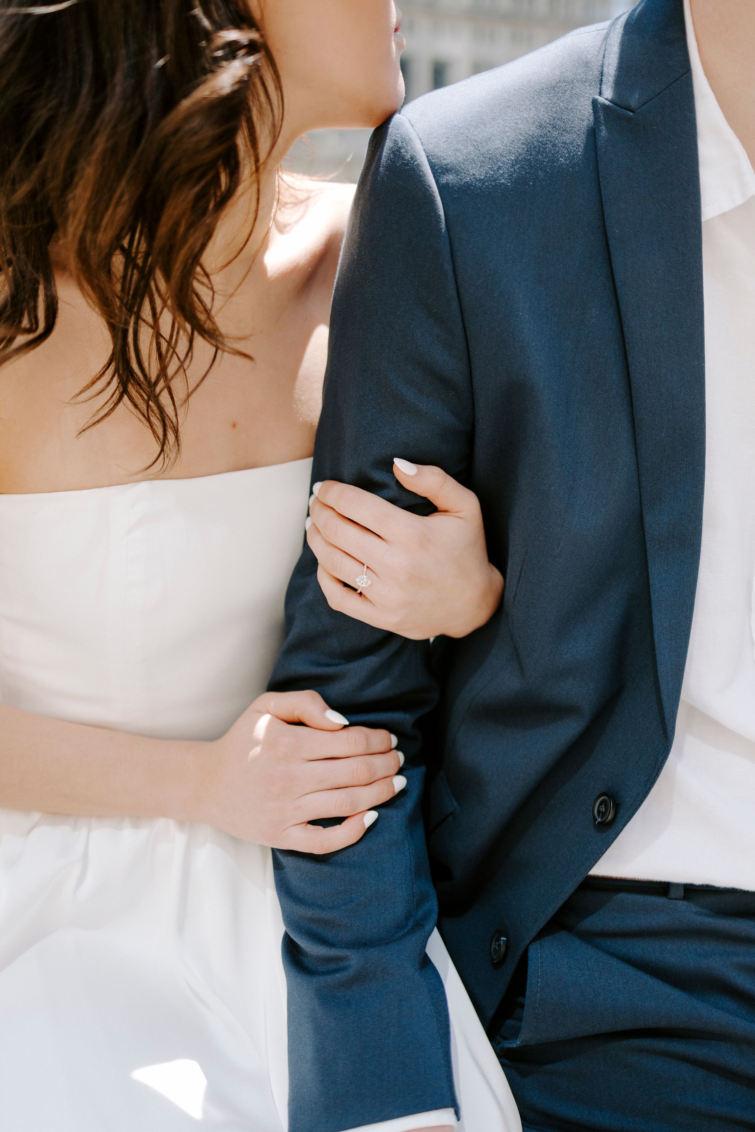 Close-up details of a couple embracing, showing a navy suit jacket and white dress in soft natural light.