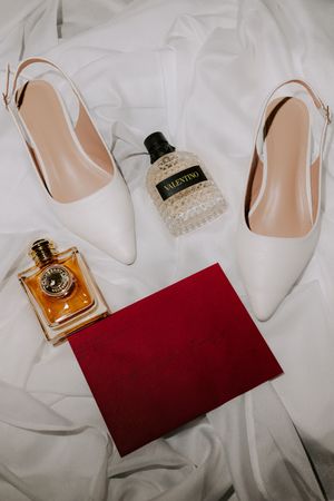 White bridal heels, Valentino perfume, amber fragrance bottle, and red velvet clutch arranged on white silk fabric.