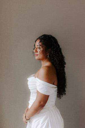 A bride in an off-shoulder white gown with curly black hair smiles softly against a neutral background.
