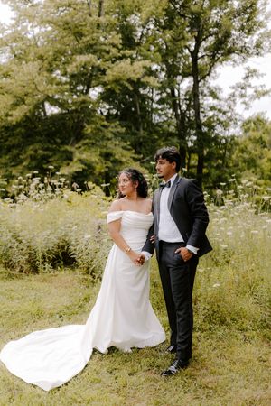 Bride in off-shoulder white gown and groom in black tuxedo pose together in a lush green garden setting.