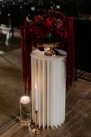 Elegant wedding decor with deep red floral arrangement in gold urn on white pedestal, candles on hardwood floor.