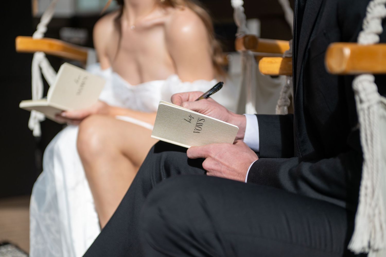 A close up image of a bride and groom sitting in hammock chairs with their vow books open. Each is writing. You cannot see their faces.