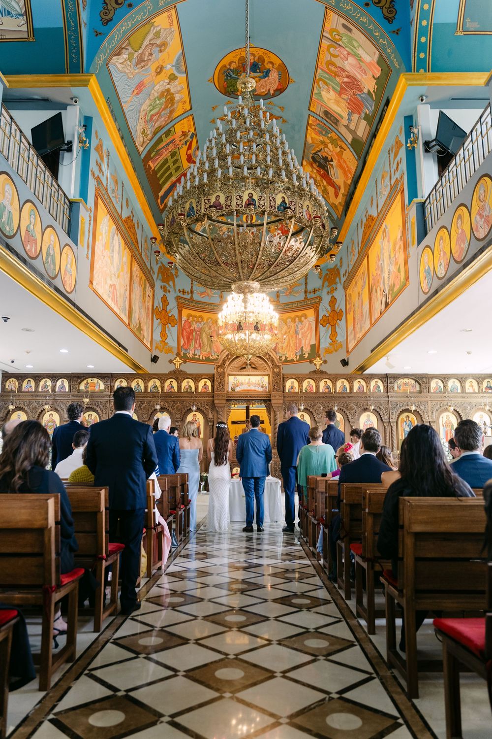 Ornate Orthodox church interior with grand chandelier, painted ceiling murals, and congregation gathered for service.