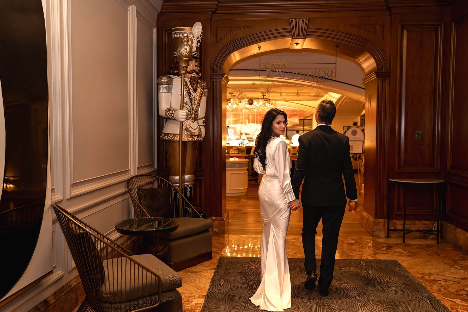 A couple in formal attire walks through an elegant arched doorway in a warmly lit historic venue.