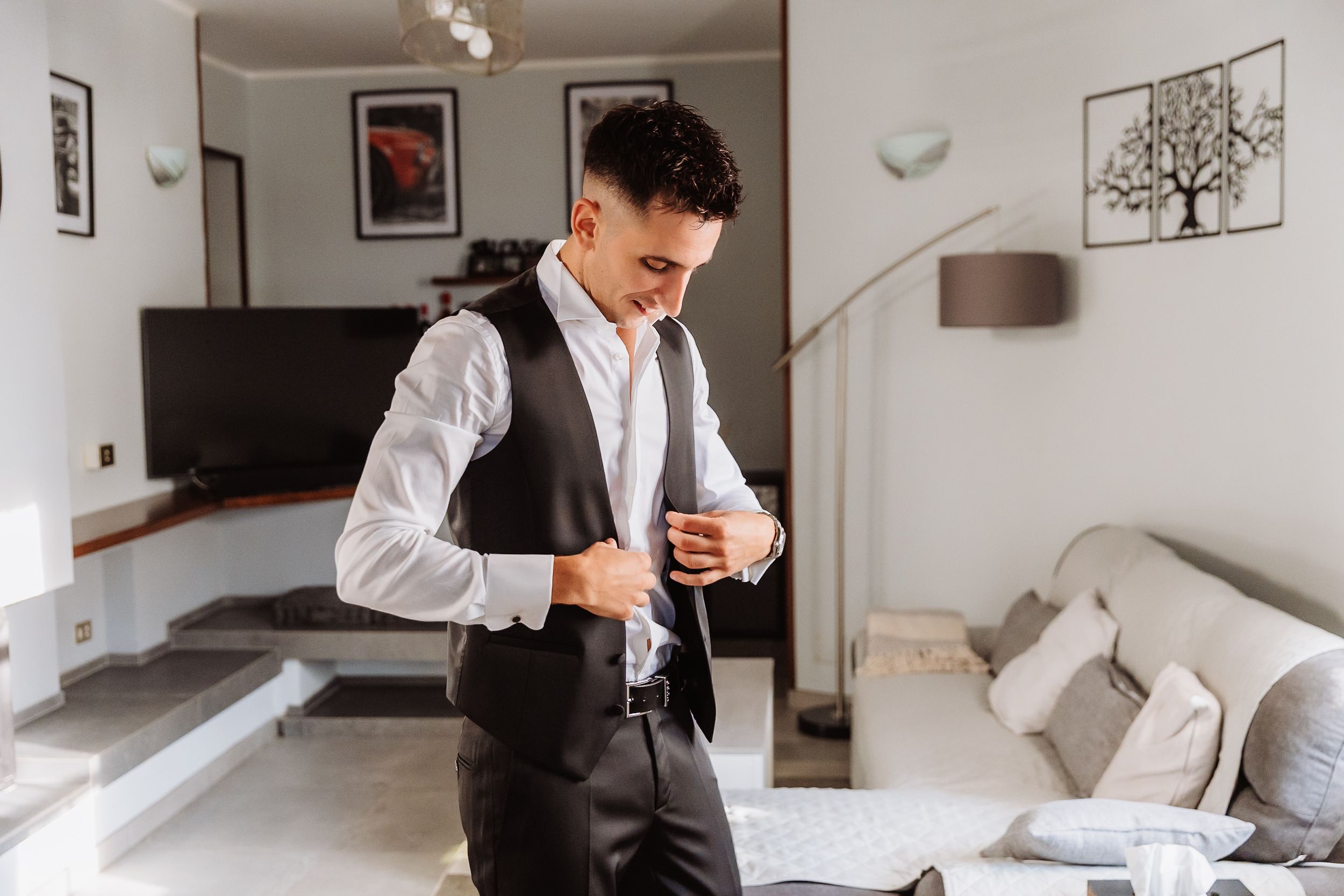 A person in formal attire with a white dress shirt and dark vest stands in a modern living room holding a drink.