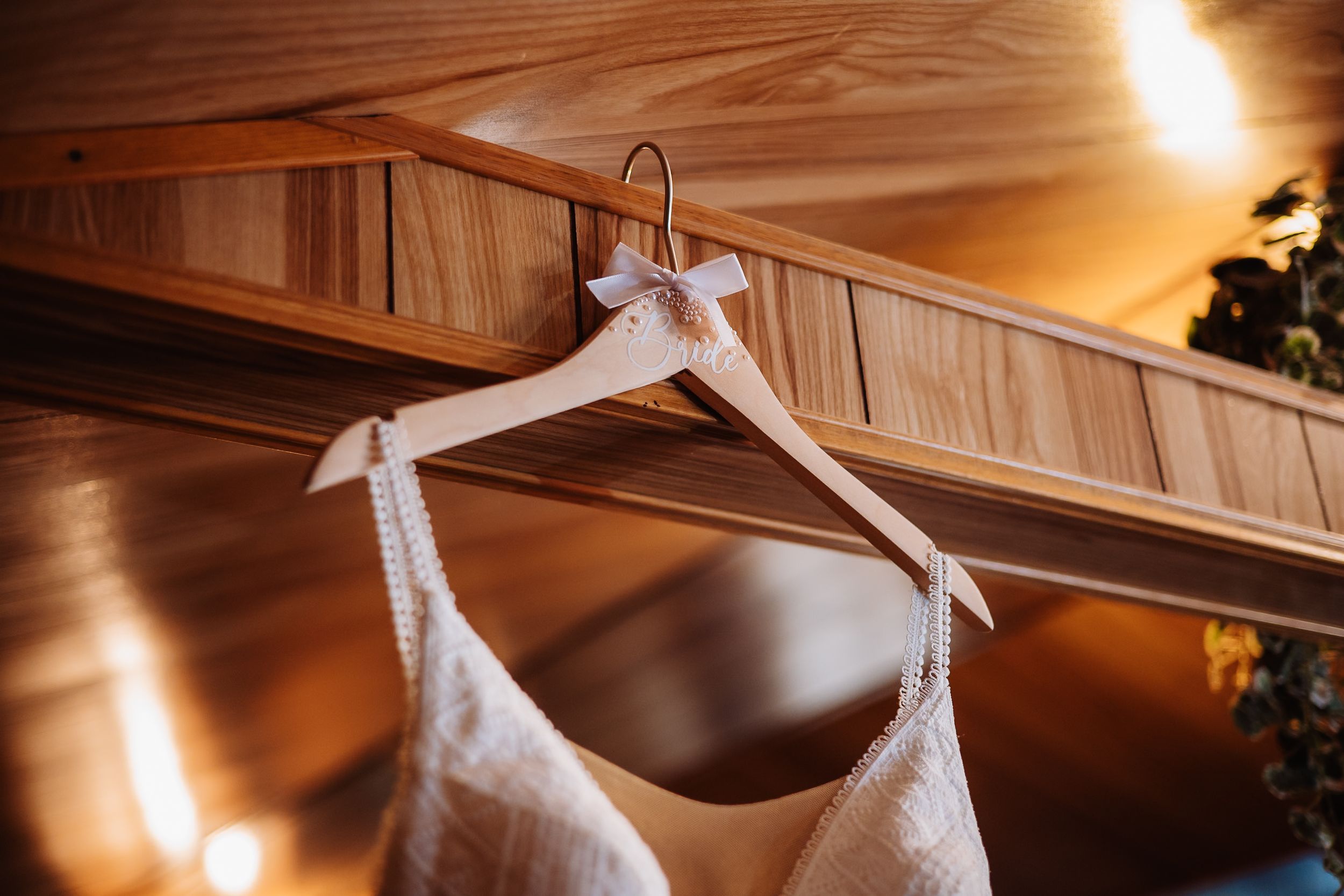 White wedding dress hangs from wooden beam in warm lighting creating an elegant bridal preparation scene.