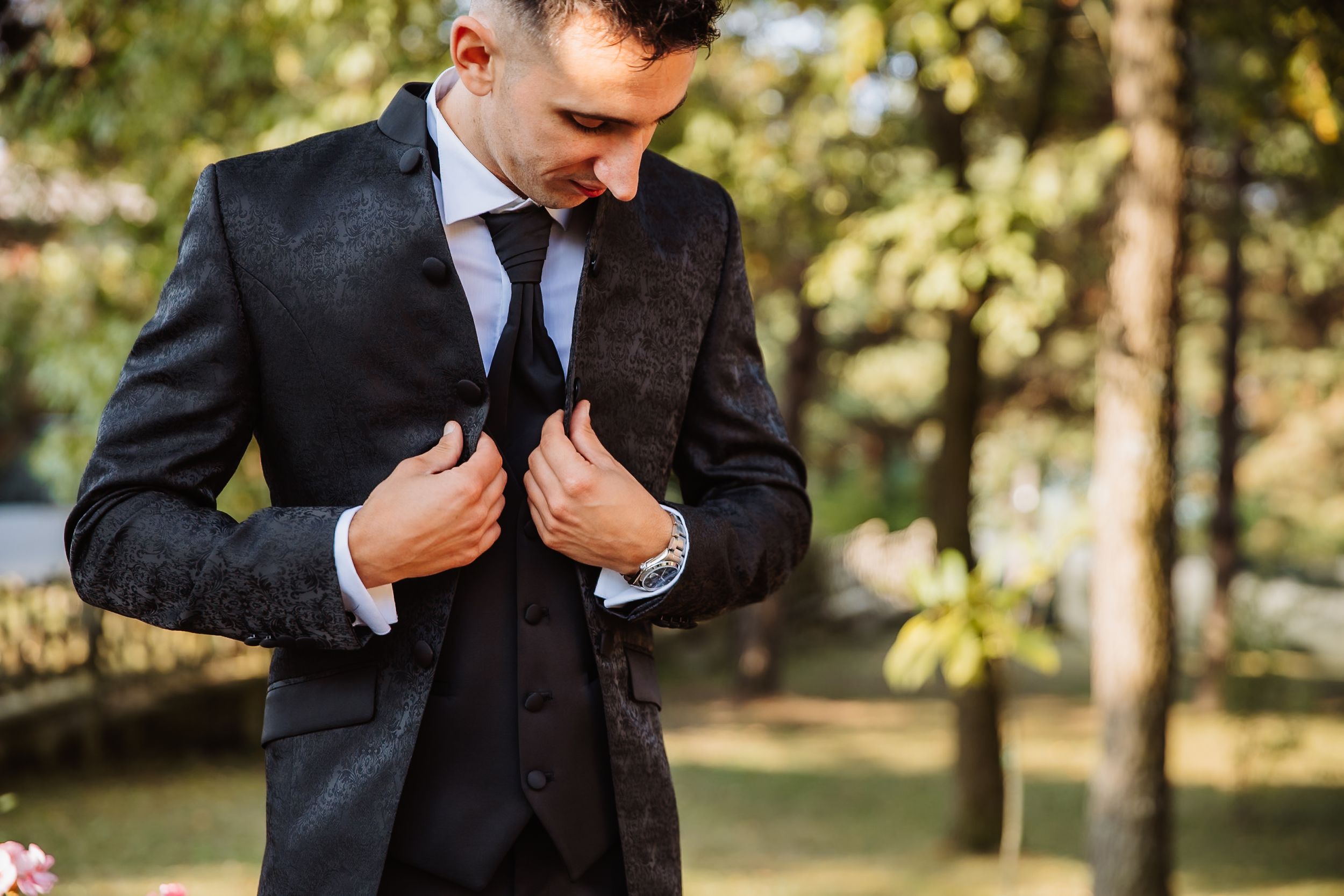 Groom adjusts his suit jacket and tie while getting ready outdoors in natural lighting.