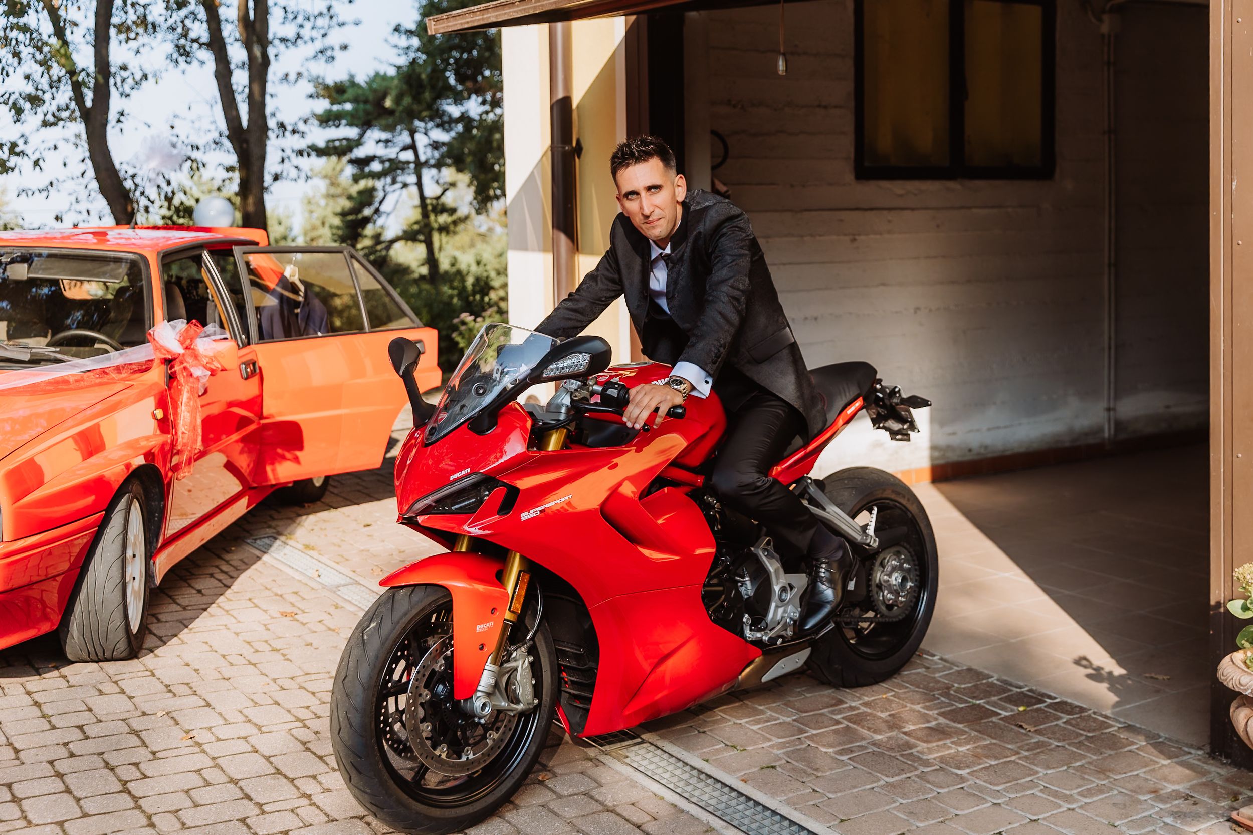 Red Ducati motorcycle parked on brick driveway with garage and trees in background.