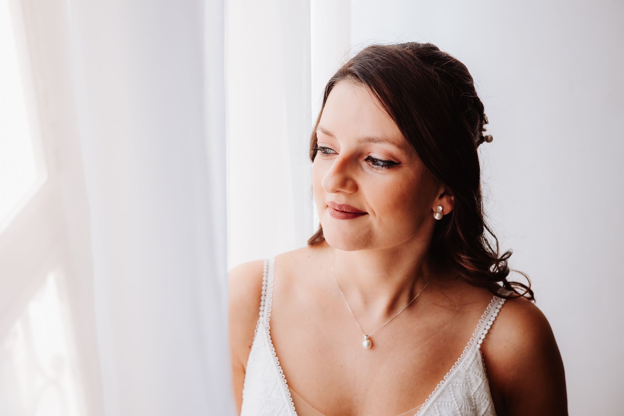A person in a white dress stands near a window with soft natural light streaming in.