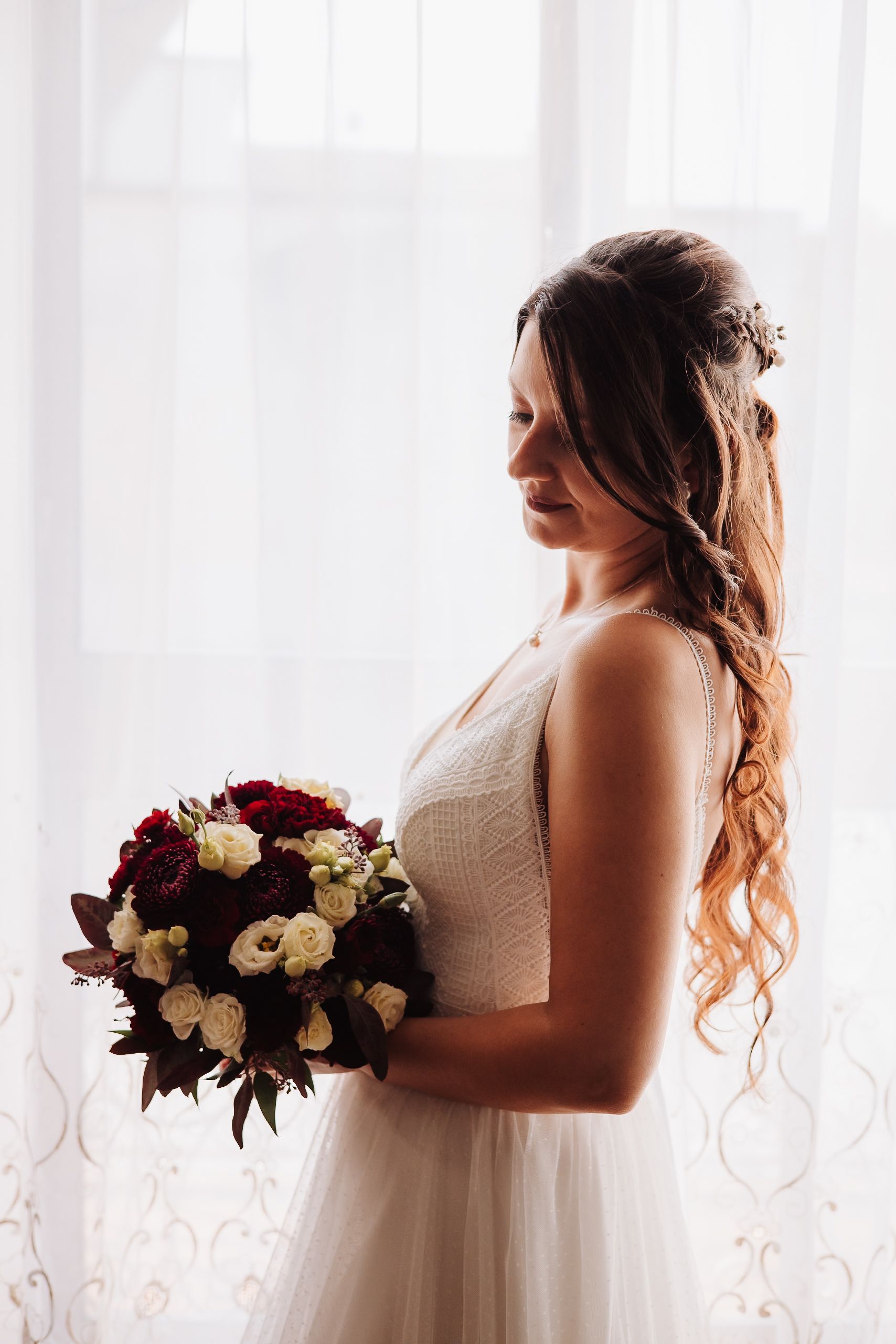 A bride in a white gown holds a burgundy and cream bouquet while posing against a bright window.