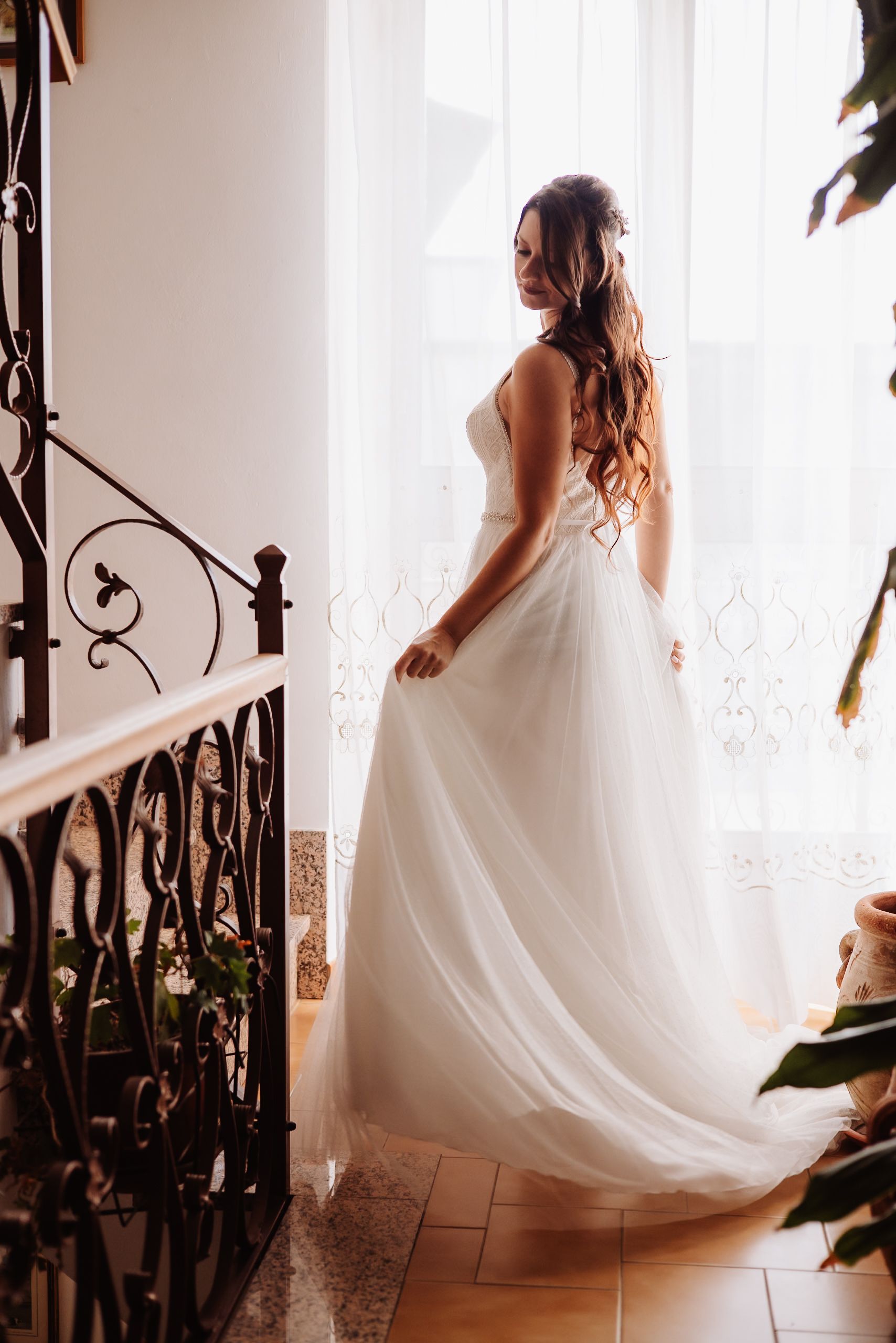 A bride in a flowing white wedding gown stands by an ornate staircase railing in soft window light.