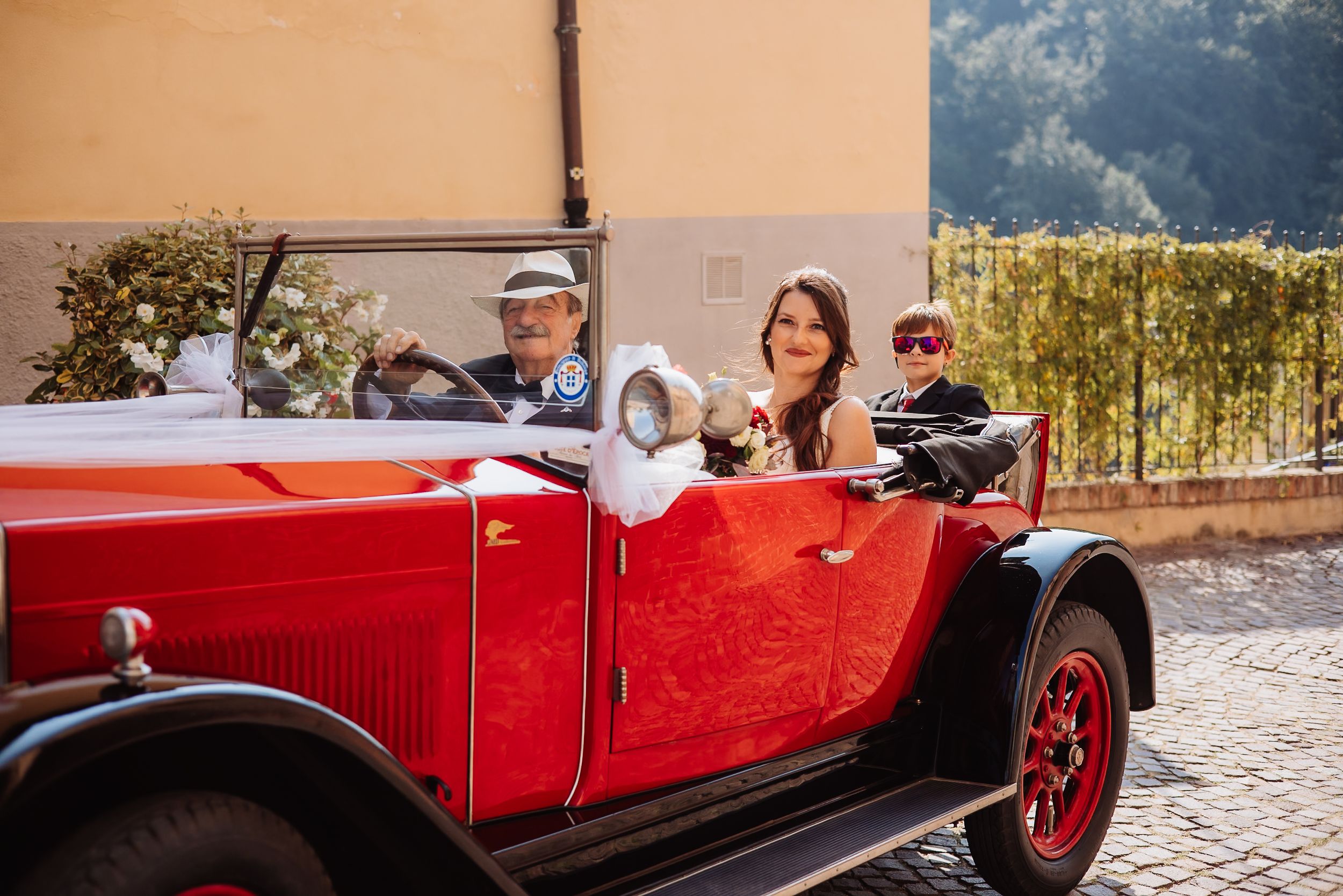 A red vintage convertible car parked on a cobblestone street with passengers enjoying a sunny day.