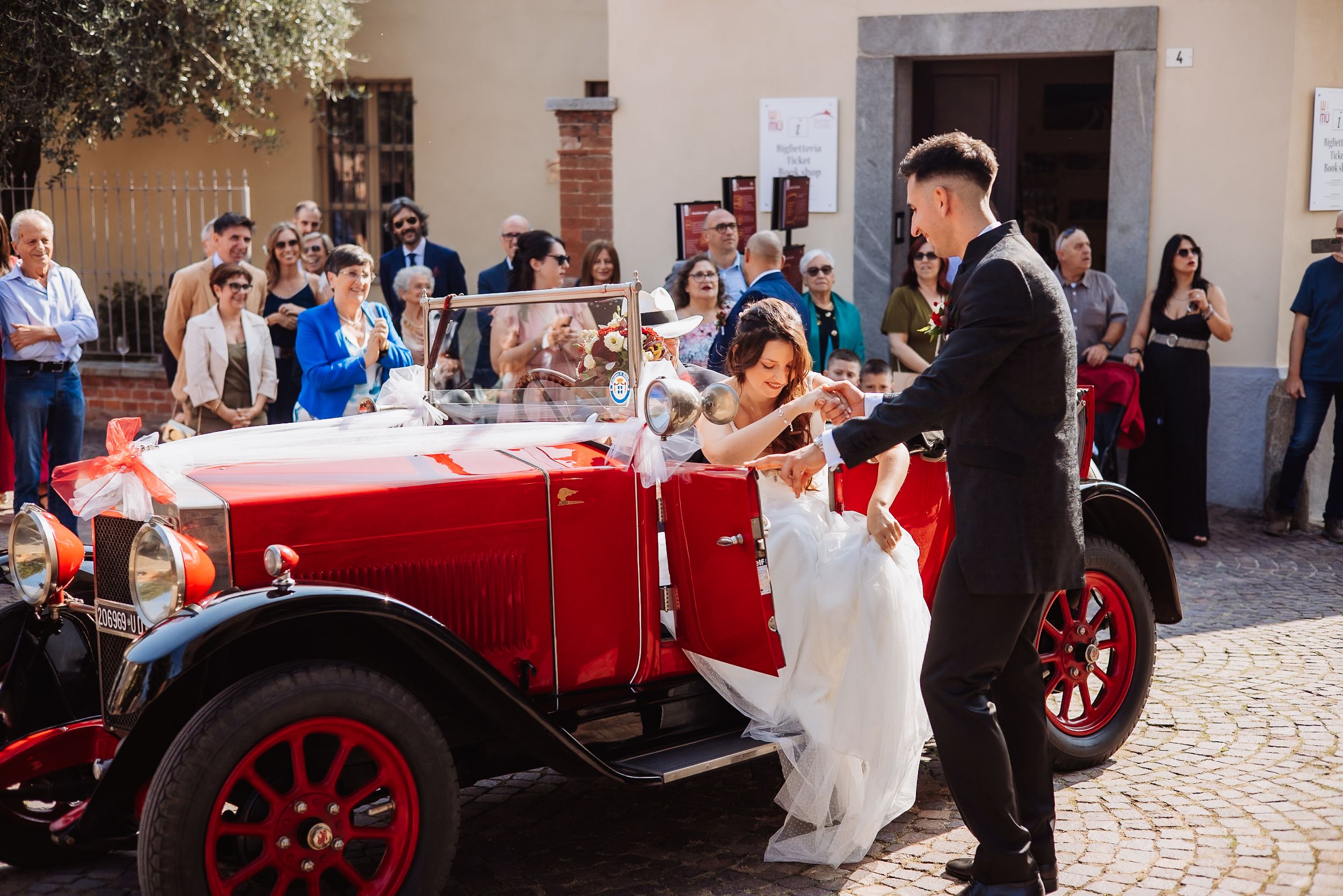 A vintage red car decorated for a wedding celebration is parked on a cobblestone street.