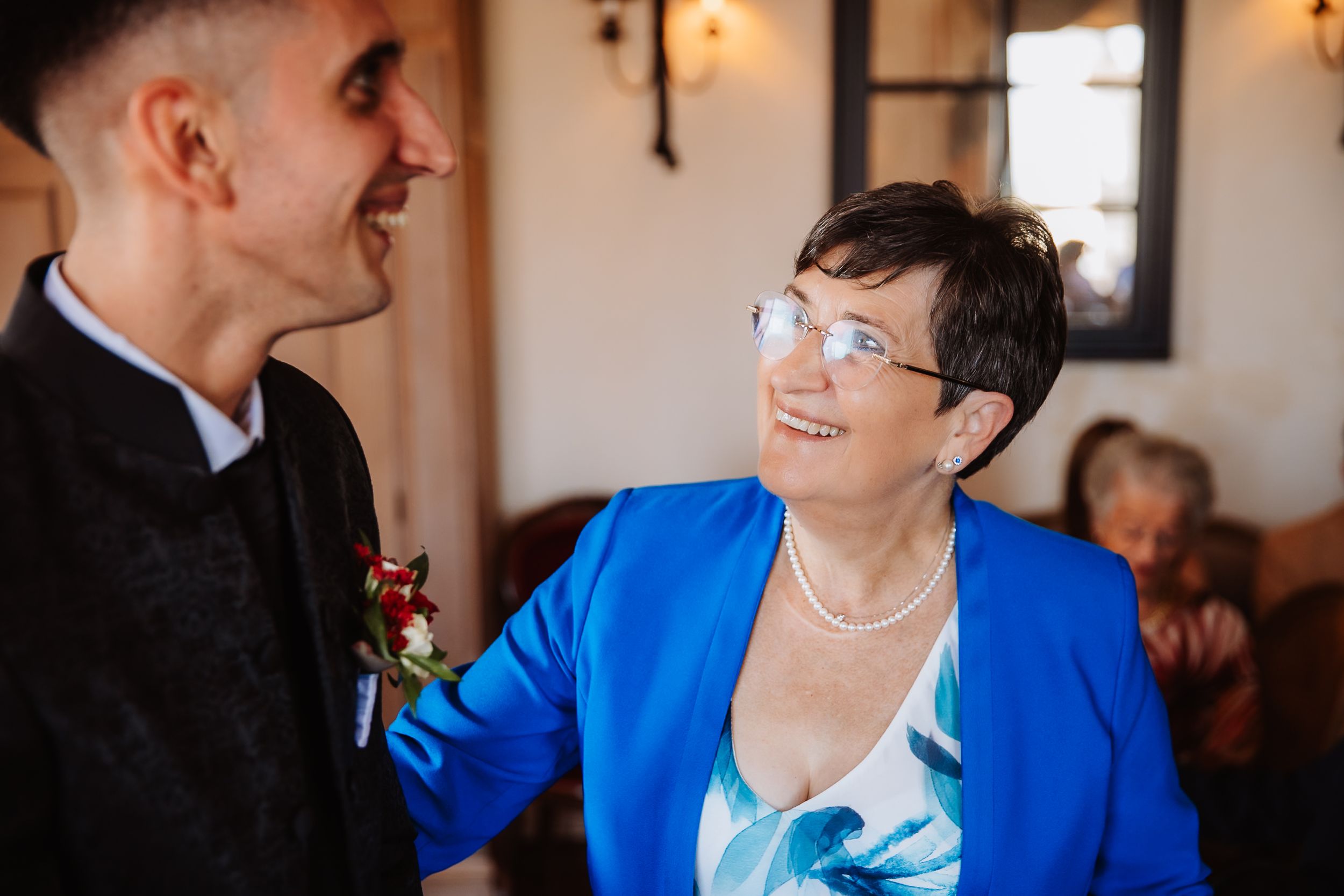 A person wearing a vibrant blue blazer smiles warmly while engaged in conversation at an indoor gathering.