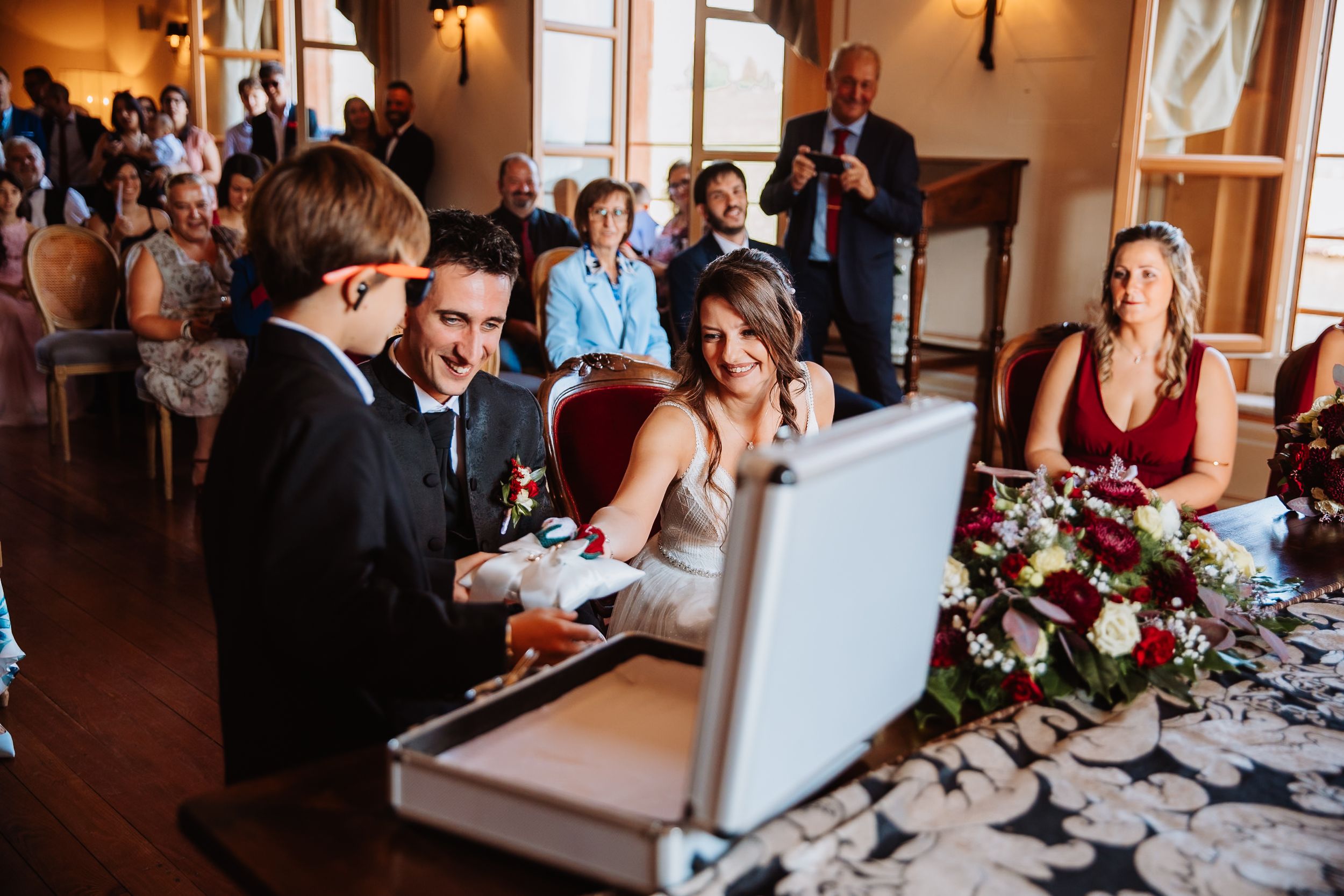 Wedding guests seated at a reception table laugh while looking at a laptop computer display during dinner.