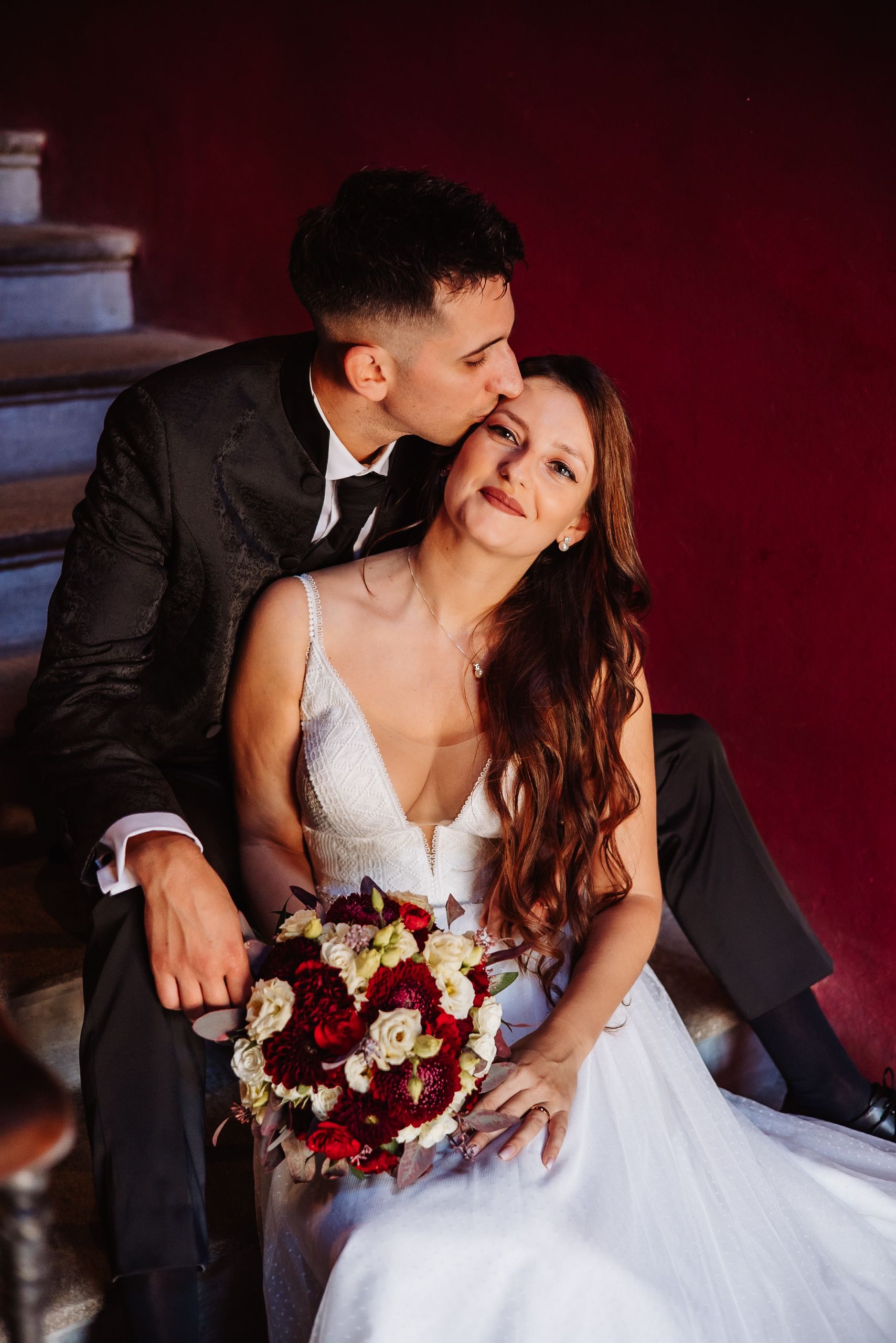 A romantic wedding couple shares intimate moments while holding a red and white floral bouquet.