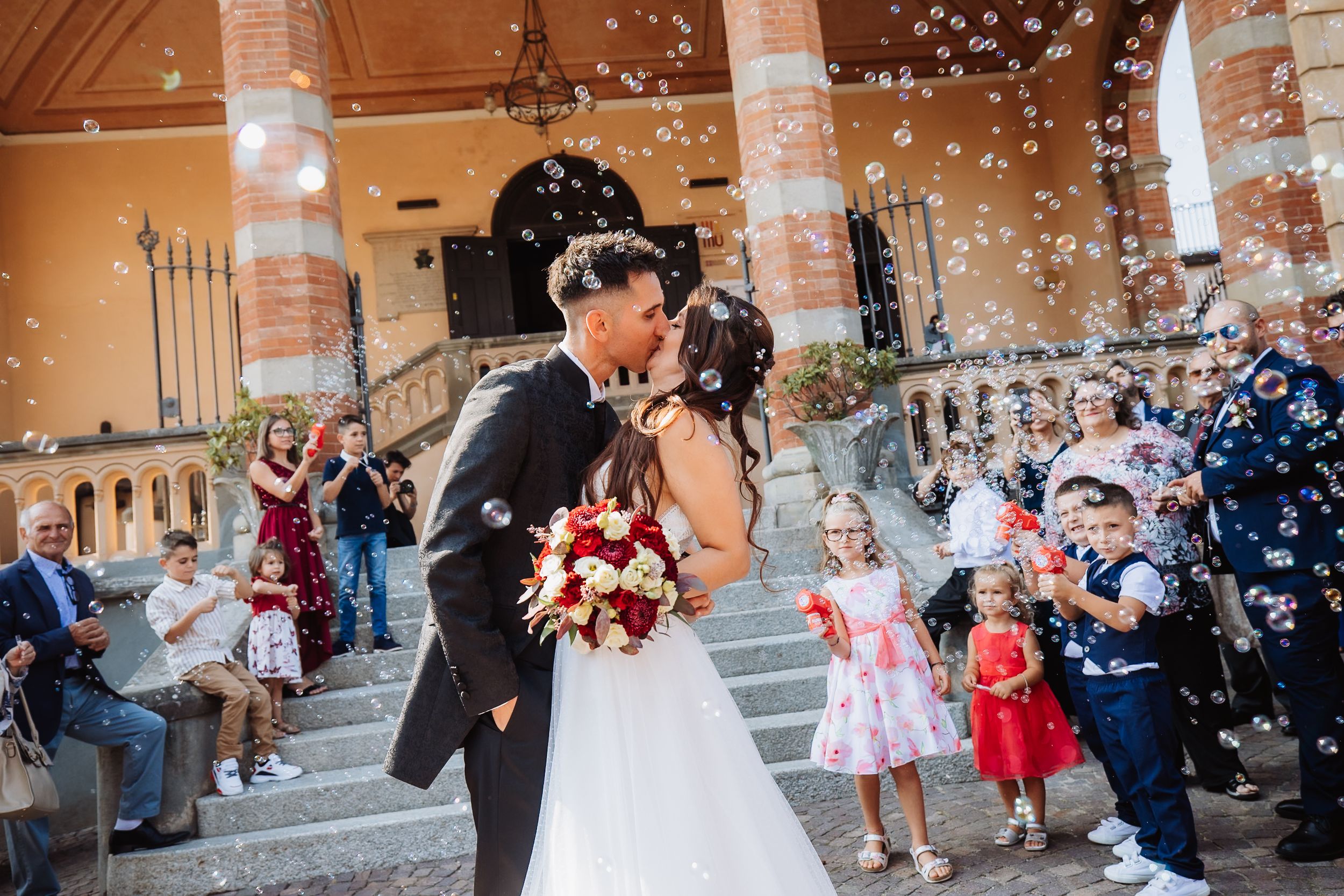 Wedding guests throw confetti as newlyweds walk through a stone archway corridor at sunset.