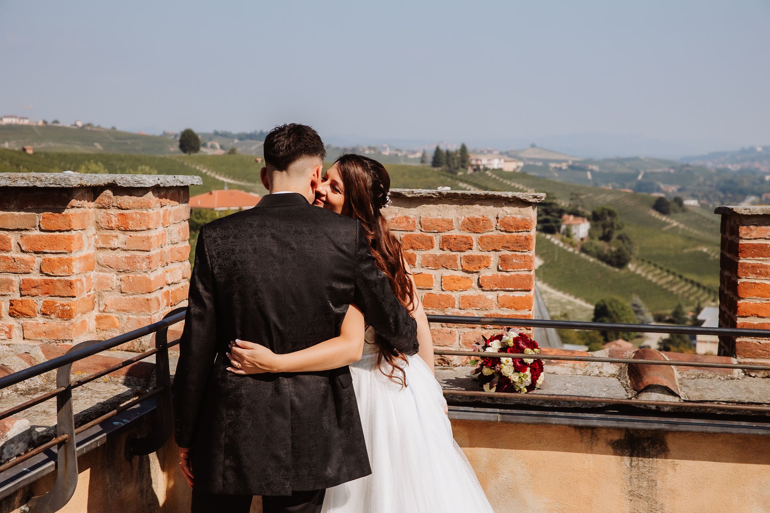 A couple shares a romantic moment on a balcony overlooking the rolling hills of an Italian countryside at sunset.
