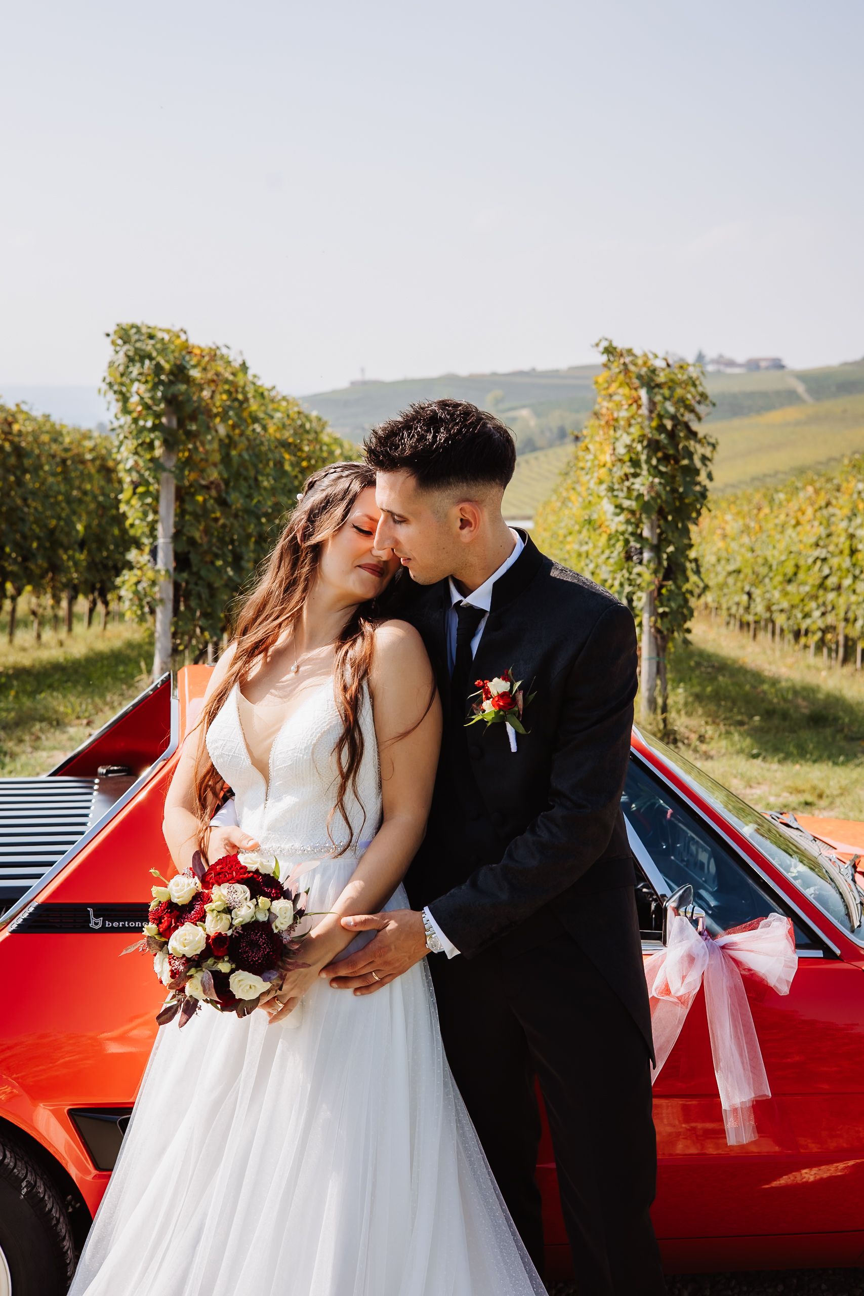 A couple shares romantic moments next to a red vintage car in a scenic vineyard setting during their wedding photoshoot.