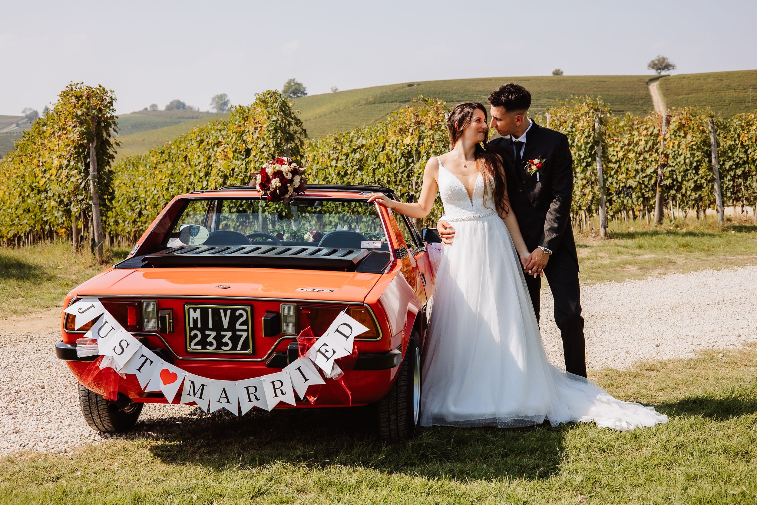 A wedding couple poses in a scenic vineyard setting with a vintage red car decorated with 'Just Married' signage.