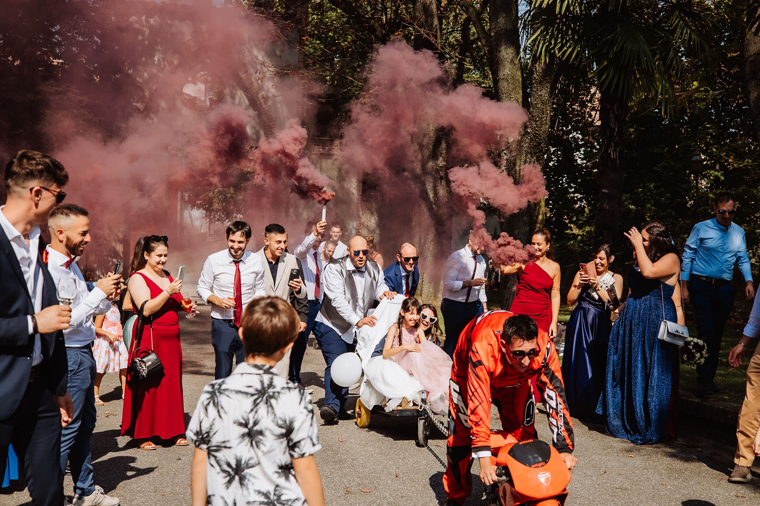 Wedding guests react with joy and emotion as pink smoke fills the outdoor ceremony space during a special moment.