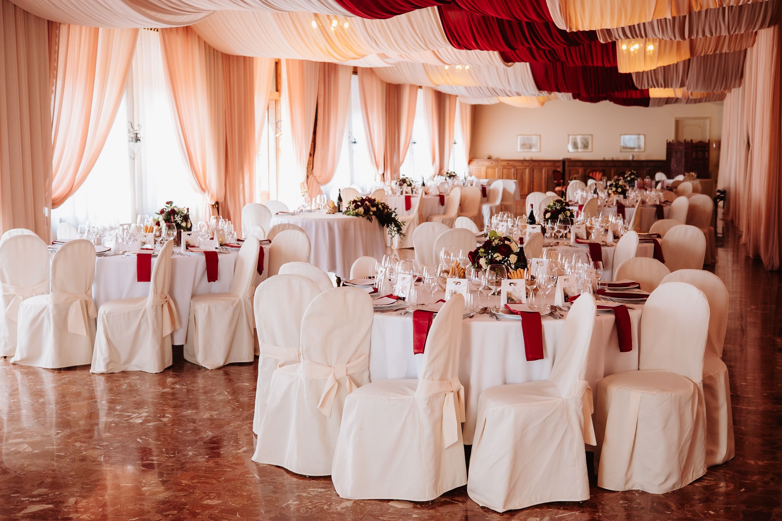 Elegant wedding reception venue with white draped fabric, chandeliers and round tables covered in white linens.