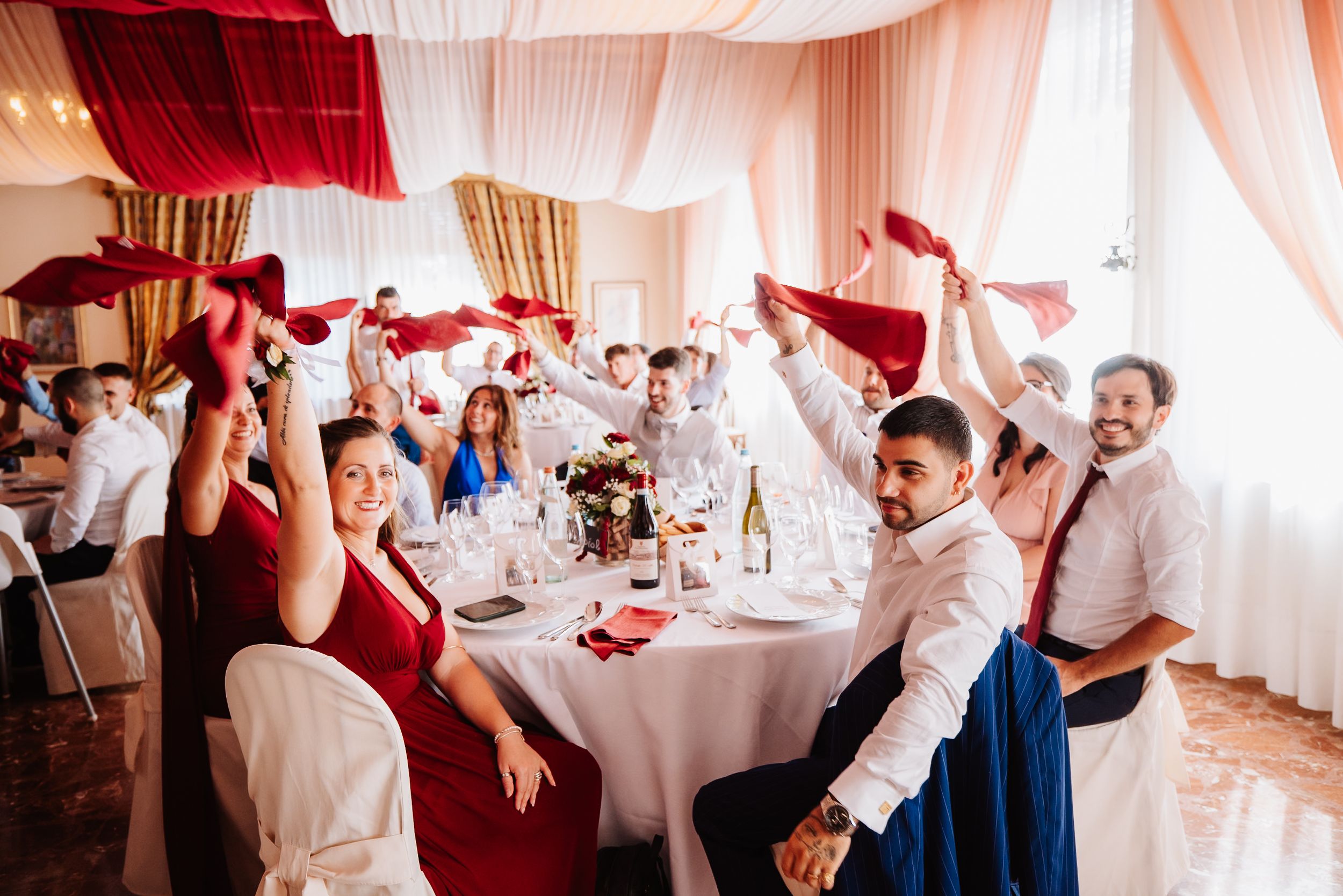 Wedding guests dancing and celebrating at an elegant reception with white drapery and red decor details.