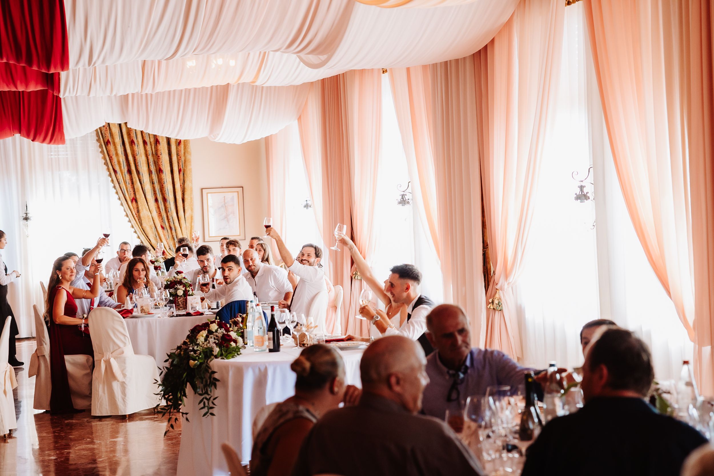 Wedding reception guests celebrate with raised glasses in an elegant white-draped ballroom during a toast.