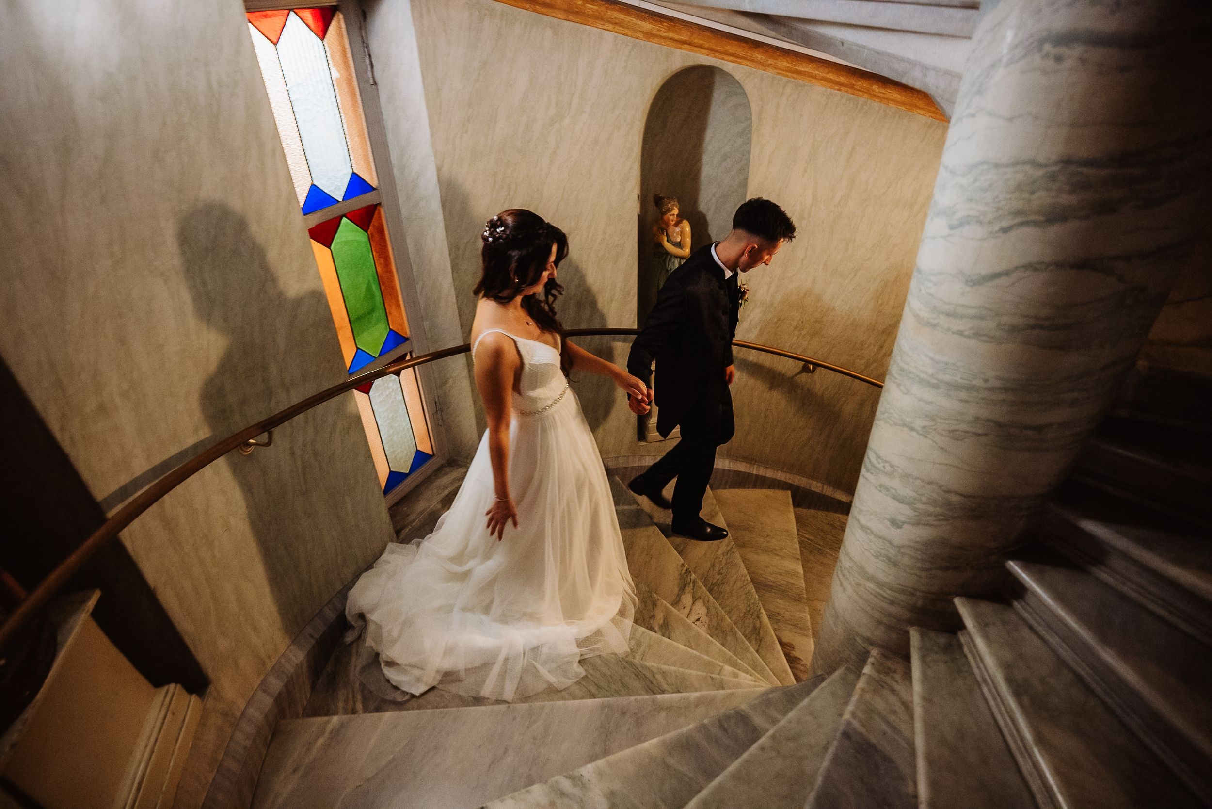 A wedding dress train flows down a curved stone staircase as people descend in an elegant indoor setting.