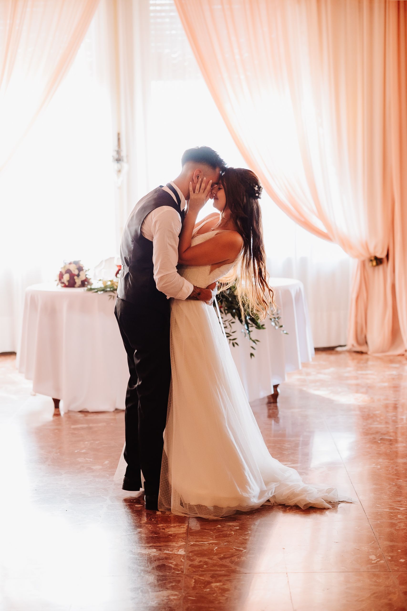 A romantic wedding couple shares their first kiss in an elegant white-draped reception venue with soft lighting.