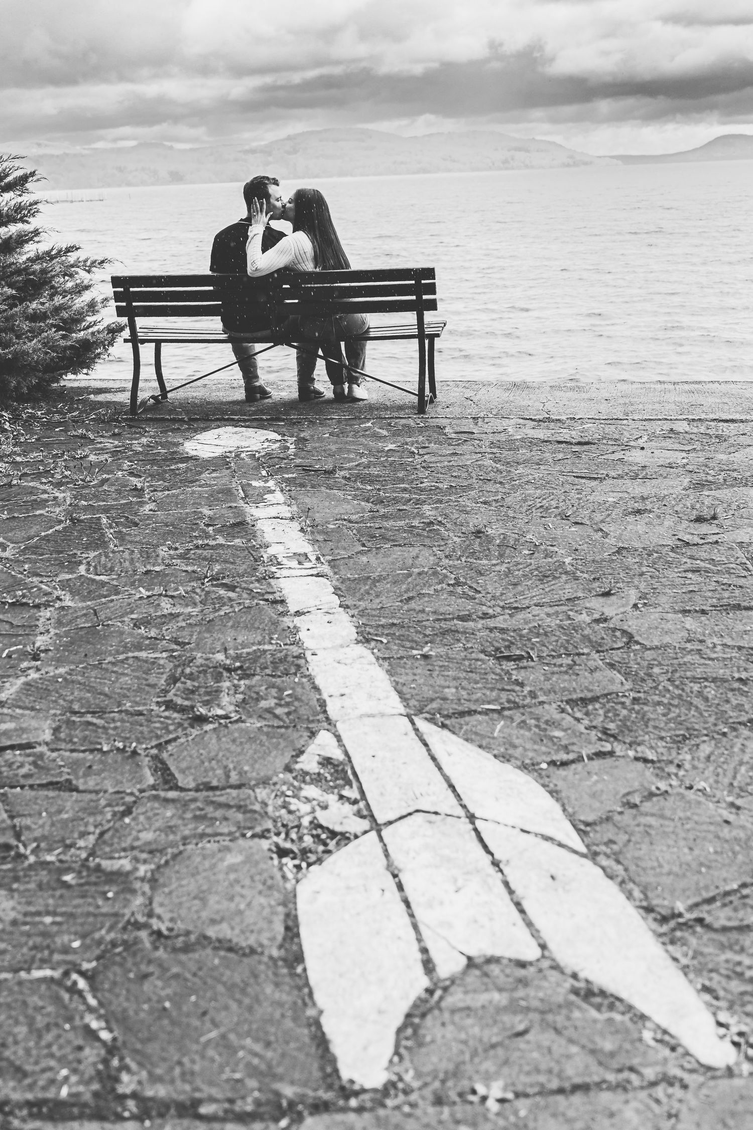 Black and white romantic photo of couple sitting on bench by the sea with arrow on cobblestone path.