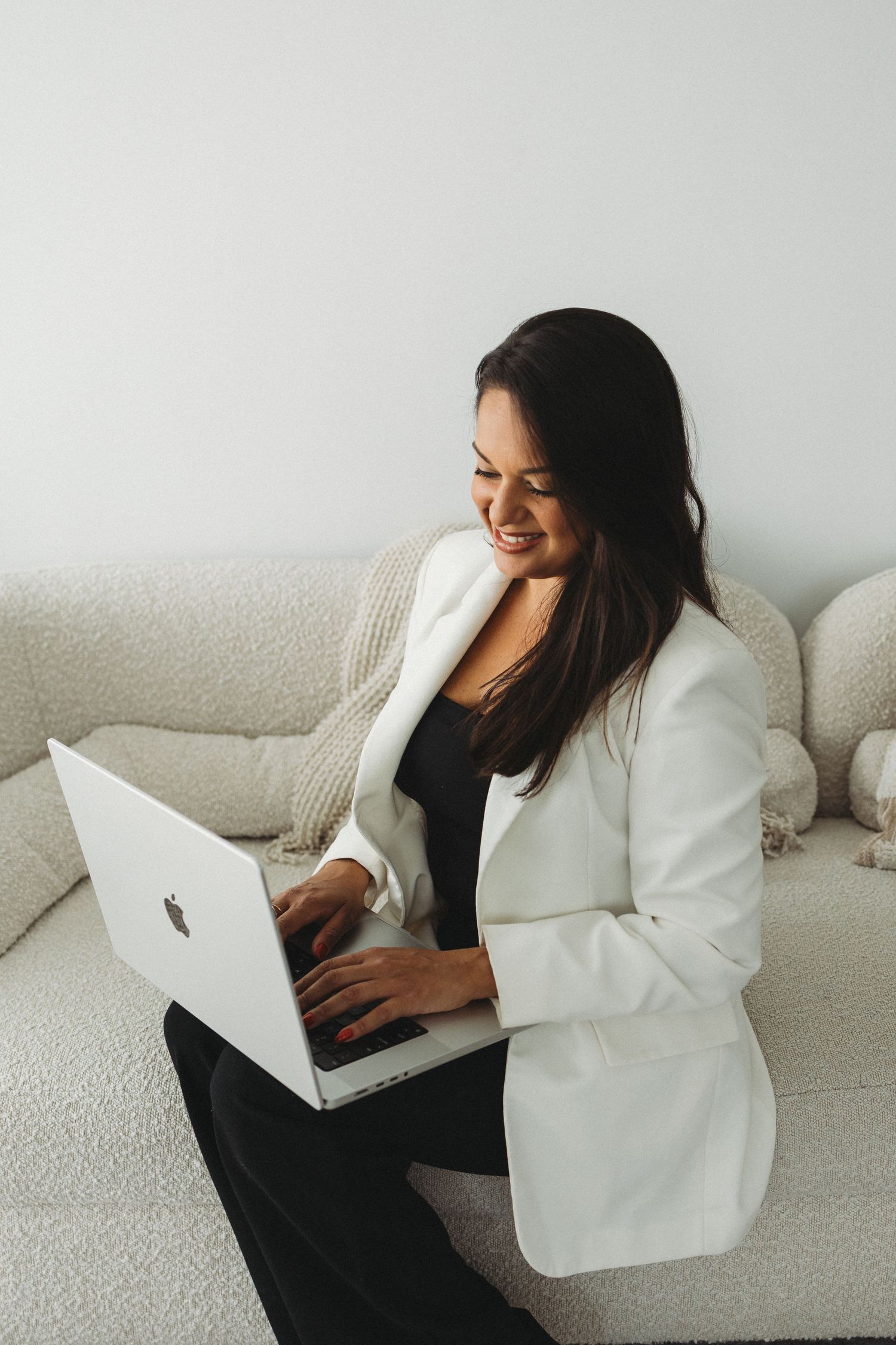 Woman in white blazer works on laptop while relaxing on light gray couch.
