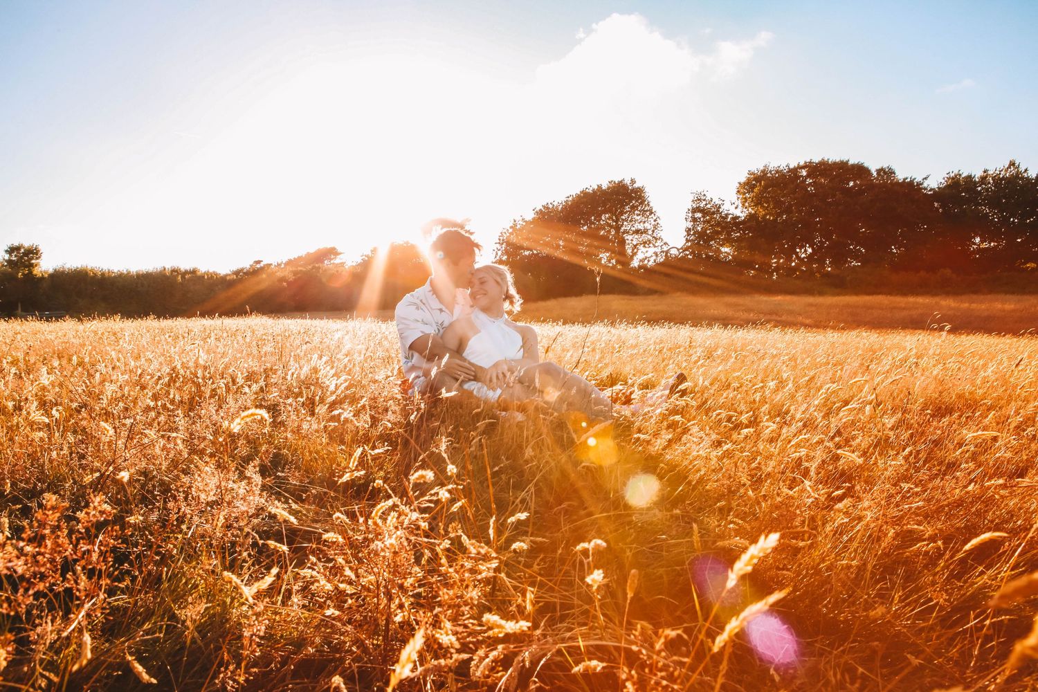 Family playing and lifting child in sunny meadow with tall grass and trees.
