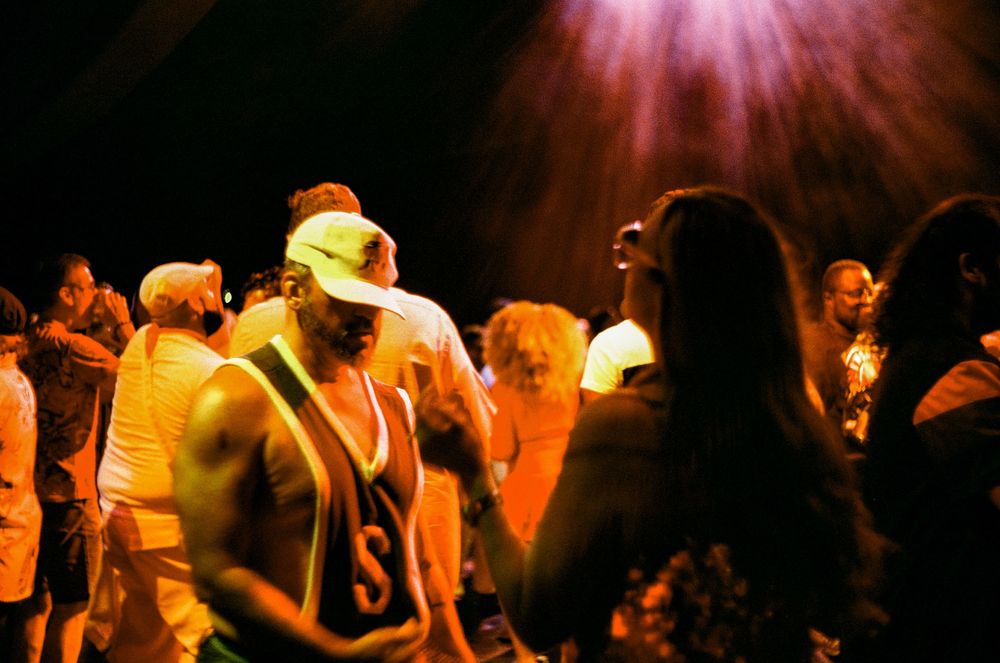 Illuminated concert crowd dancing with raised hands silhouetted against bright stage lights and pink glow.