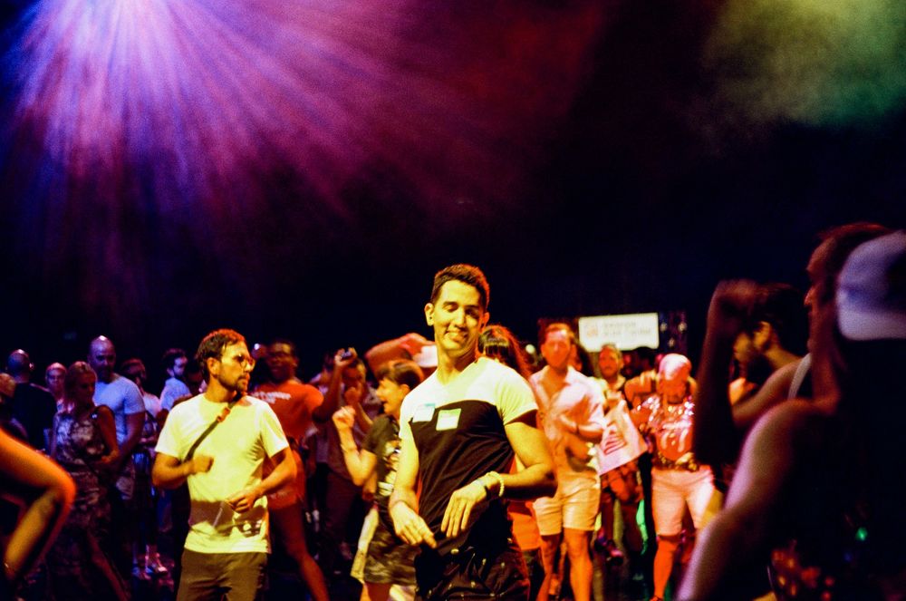 A crowded dance hall filled with people moving under dramatic purple and blue stage lighting.