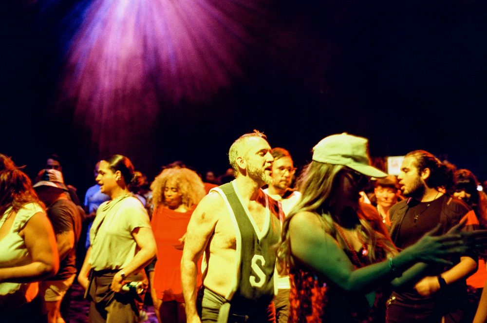 Crowded dance floor with people dancing under vibrant purple and yellow lighting at a nightclub or party.