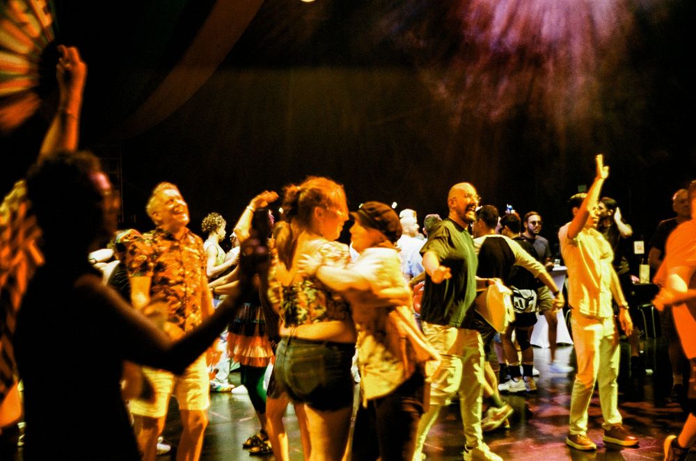 Performers in colorful outfits dance energetically on stage under vibrant theatrical lighting during a show.
