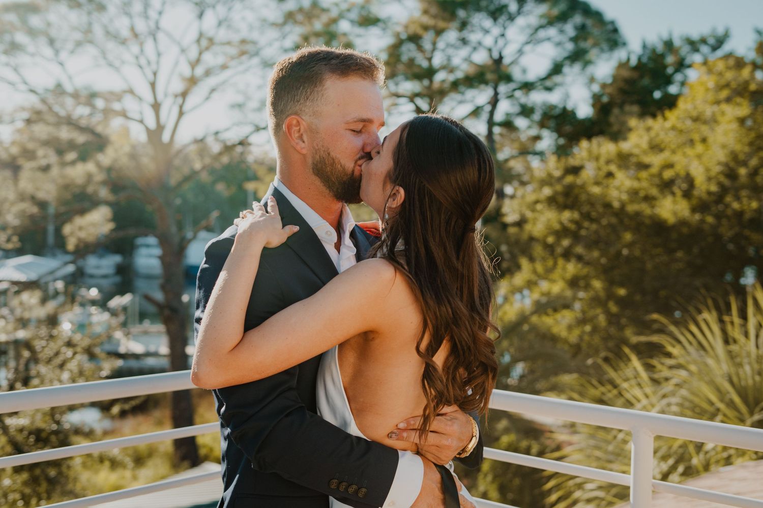 A couple shares an intimate kiss on an outdoor deck surrounded by lush greenery and sunlit trees in the background.